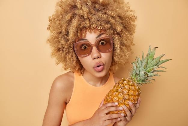 Porträt der überraschten Frau hält die Lippen abgerundet sieht mit Erstaunen hält Ananas trägt trendige Sonnenbrillen und lässiges T-Shirt isoliert über beige Hintergrund Weibliches Modell mit exotischen Früchten