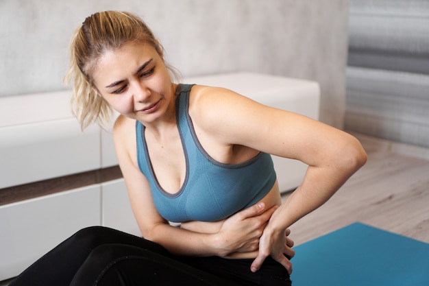 Porträt der unglücklichen jungen Frau, die auf Yogamatte sitzt, ihren Rücken nach dem Training berührt, unter Rückenschmerzen leidet, Schmerzen fühlt, Seitenansicht