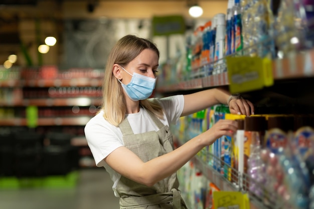 Porträt der Verkäuferin, in schützender Gesichtsmaske, die im Supermarkt lächelt und in die Kamera schaut. Angenehme freundliche Verkäuferin, die im Laden zwischen den Reihen steht. Arbeiter mit verschränkten Armen