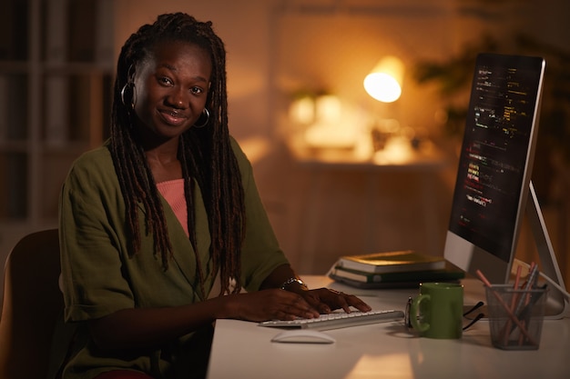 Porträt der zeitgenössischen afroamerikanischen Frau, die Code schreibt und Kamera beim Arbeiten im dunklen Büro, Kopierraum betrachtet