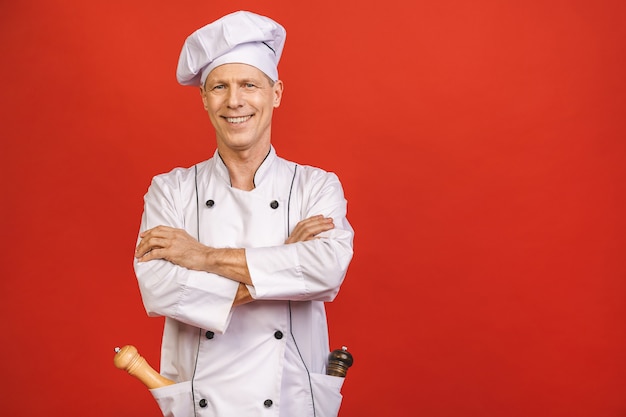 Porträt des älteren lächelnden jungen Küchenchefs, der mit Pfeffermühle lokalisiert auf rotem Hintergrund kocht.