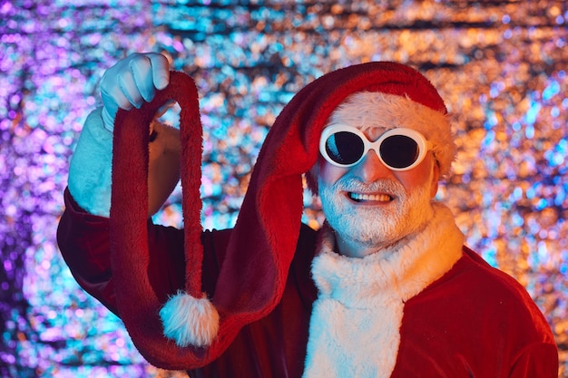 Porträt des älteren Mannes im Weihnachtsmannkostüm, das Sonnenbrille und lustigen Hut trägt, der gegen den glänzenden Hintergrund lächelt