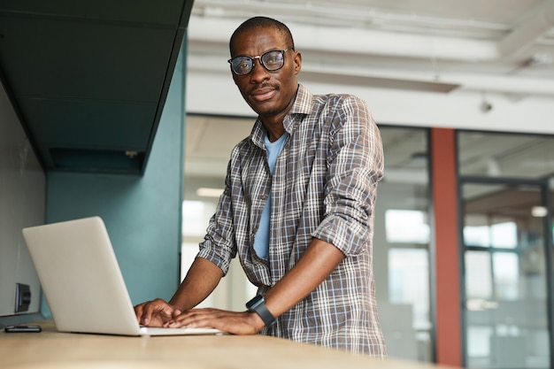 Porträt des afrikanischen Designers in der Brille, während er am Tisch steht und seinen Laptop benutzt