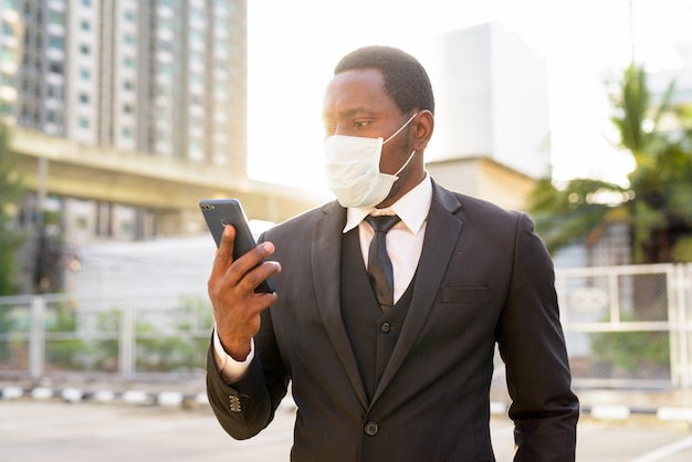 Porträt des afrikanischen Geschäftsmannes mit Maske unter Verwendung des Telefons in den Stadtstraßen