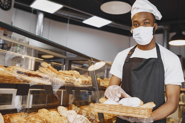 Porträt des afroamerikanischen Bäckers mit frischem Brot in der Bäckerei. Konditor, der kleines Gebäck hält.