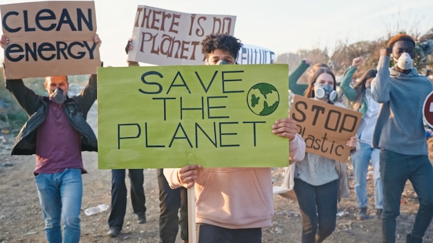 Porträt des Aktivisten des jungen Mannes mit einem Plakat, das dazu aufruft, sich um die Umwelt zu kümmern. Im Hintergrund kämpfende Menschen, die gegen die Verschmutzung durch Müll protestieren, die auf einer Müllkippe am Stadtrand bleiben.