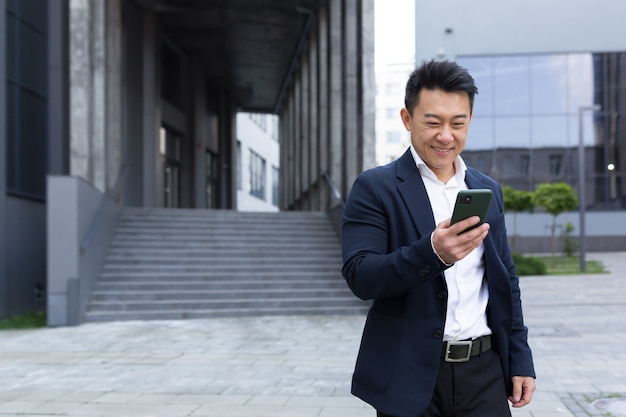 Porträt des asiatischen fröhlichen und erfolgreichen Geschäftsmannes in der Nähe des Büros im Anzug bekam gute Nachrichten, gute Nachrichten online gelesen