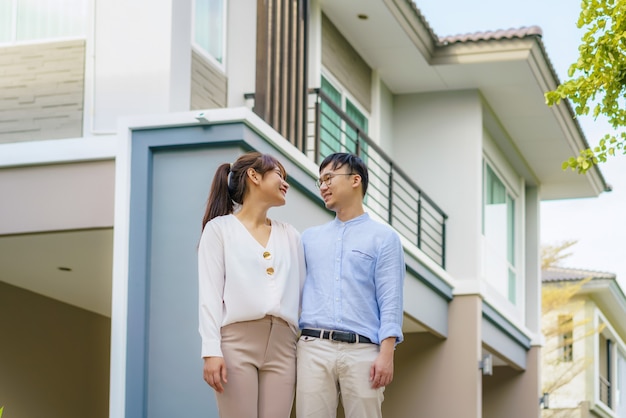 Porträt des asiatischen jungen Paares, das zusammen steht und sich umarmt und glücklich vor ihrem neuen Haus schaut, um neues Leben zu beginnen. Familien-, Alters-, Haus-, Immobilien- und Personenkonzept.