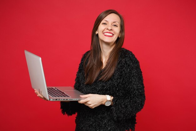 Porträt des atemberaubenden lächelnden jungen Mädchens im schwarzen Pelzpullover, der Laptop-PC-Computer lokalisiert auf hellem rotem Wandhintergrund im Studio hält. Menschen aufrichtige Emotionen Lifestyle-Konzept. Kopieren Sie Platz.