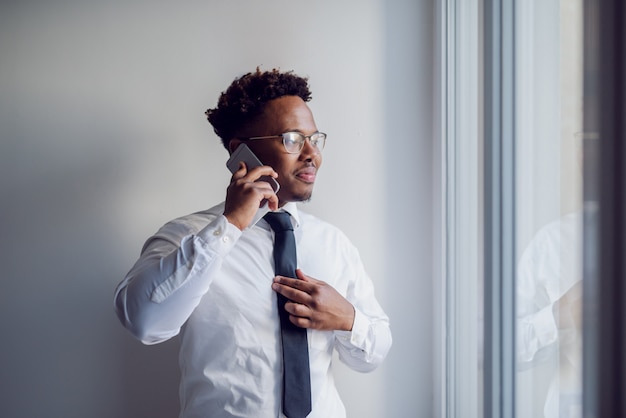 Porträt des attraktiven afroamerikanischen Geschäftsmannes, der neben Fenster steht und durch ihn schaut, während er ein Telefongespräch mit Kollege hat.