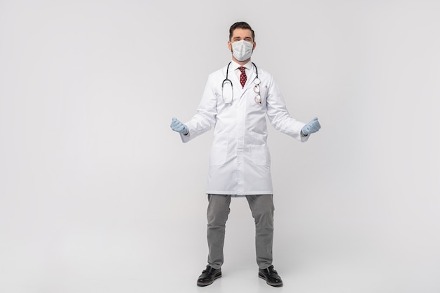 Porträt des attraktiven gutaussehenden Arztes in der schützenden Gesichtsmaske, im weißen Laborkittel, Krawatte lokalisiert auf weißer Wand