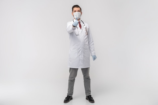 Porträt des attraktiven gutaussehenden Arztes in der schützenden Gesichtsmaske, im weißen Laborkittel, Krawatte lokalisiert auf weißer Wand
