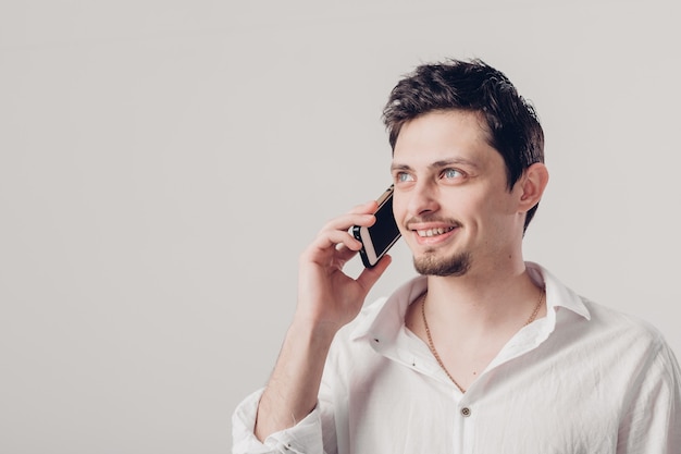 Porträt des attraktiven jungen Brunettemannes im weißen Hemd, das auf dem Smartphone auf dem grauen Hintergrund spricht. Sanftes Licht