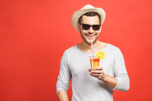 Porträt des attraktiven jungen Mannes im Hut und in der Sonnenbrille, die Orangensaft über rotem Hintergrund stehen und trinken