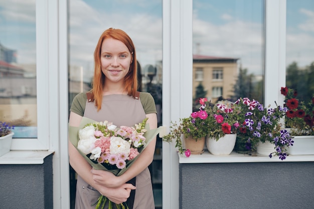 Porträt des attraktiven Mädchens mit den roten Haaren, die am Balkon mit Topfblumen auf Fensterbank stehen und netten Blumenstrauß umarmen