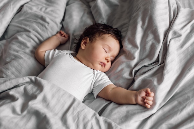 Porträt des Babys in Nachtwäsche, das zu Hause unter der Decke auf dem Bett liegt Schlafende Hände auseinander Säuglingskind im Schlafzimmer siehe süßer Traum Grauer Hintergrund, kostenloser Kopierbereich Kinderbetreuung und gesunder Schlaf-Konzept