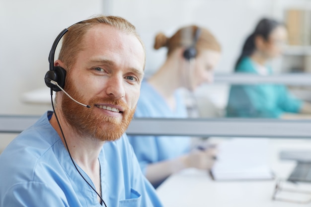 Porträt des bärtigen jungen Mannes in den Kopfhörern, die lächeln, während sie im Callcenter im Büro arbeiten
