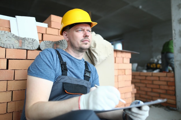 Porträt des besorgten Mannes weg schauend mit Nachdenklichkeit. Konstruktor, der Planordner hält und etwas darin aufschreibt. Bärtiger Spezialist, der an Arbeitsplan denkt. Gebäudekonzept