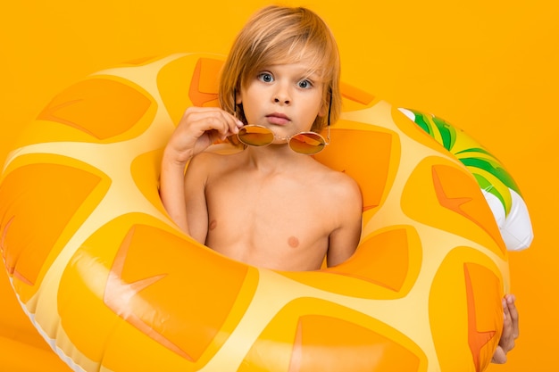 Porträt des blonden Jungen in der gelben Badehose und in der Sonnenbrille mit einem fadenscheinigen Schwimmkreis der Ananas auf Orange