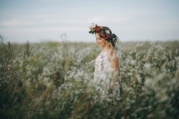 Porträt des blonden Mädchens mit blauen Augen mit einem Blumenkranz auf ihrem Kopf, der im Feld mit weißen Blumen geht