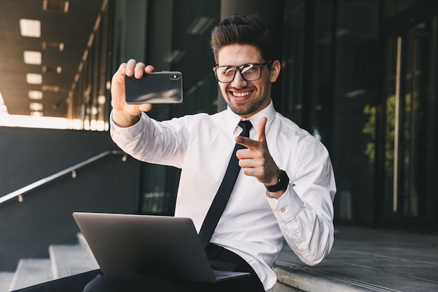 Porträt des erfolgreichen Geschäftsmannes gekleidet im formellen Anzug, der außerhalb des Glasgebäudes mit Laptop sitzt und Selfie-Foto auf Handy nimmt