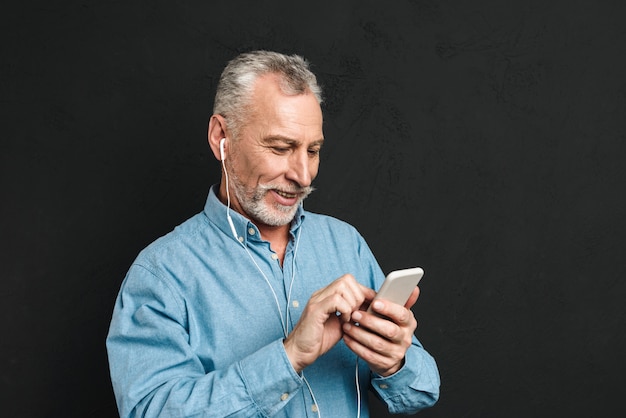 Porträt des erfreuten männlichen Rentners 60s mit grauem Haar, das auf Smartphone chattet oder im Internet surft, lokalisiert über schwarzer Wand