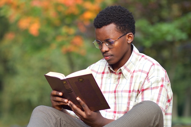 Porträt des ernsthaften konzentrierten jungen afroamerikanischen intelligenten Mannstudenten in den Gläsern und im Hemd, die ein Buch im goldenen Herbstpark lesen