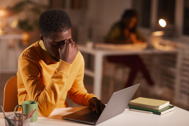 Porträt des erschöpften afroamerikanischen Mannes, der die Augen reibt, während er spät in der Nacht im dunklen Büro arbeitet, kopieren Raum