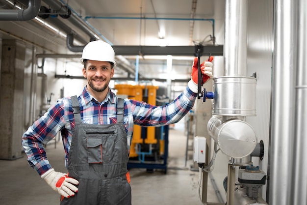 Porträt des Fabrikingenieurarbeiters, der seinen Arm auf dem Ventil hält und an Heiz- oder Kühlrohrleitungssystem im Kesselraum bereitsteht