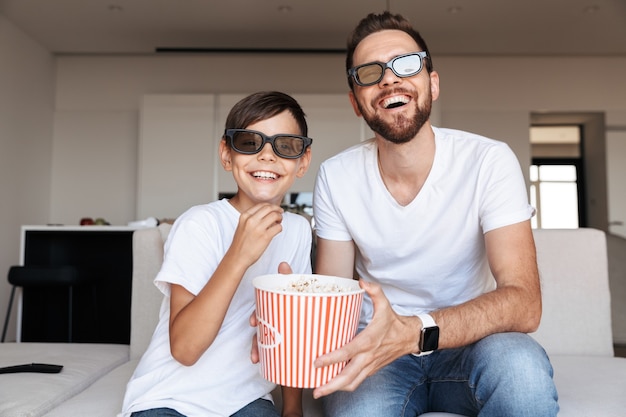 Porträt des freudigen Vaters und des Sohnes, die eine 3D-Brille tragen, die Popcorn isst und lächelt, während auf der Couch drinnen sitzen und Film schauen