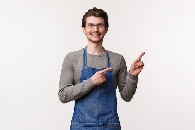 Porträt des freundlichen lächelnden Barista, des männlichen Unternehmers, der eigenes kleines Unternehmen gründet, Coffeeshop, der obere rechte Ecke zeigt, lädt ein, seine besten Getränke in der Stadt zu versuchen, auf einer weißen Wand stehen