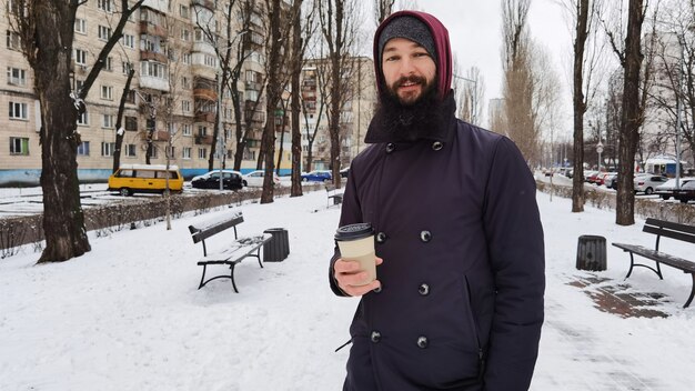 Porträt des fröhlichen attraktiven bärtigen jungen Mannes, der heißen Kaffee draußen trinkt