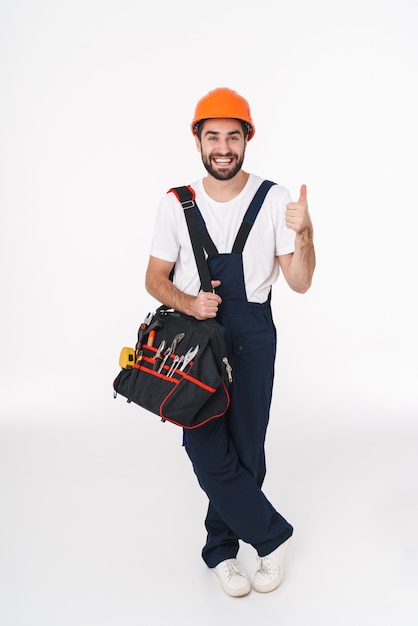 Porträt des fröhlichen jungen Baumeisters im Helm lokalisiert über der weißen Wand, die Tasche mit Ausrüstungsinstrumenten hält, die Daumen hoch Geste zeigen.