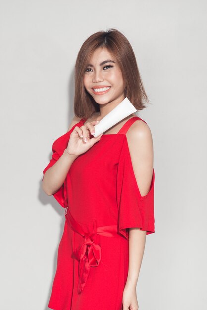 Porträt des fröhlichen lächelnden Modemädchens im roten Kleid, das kosmetisches Plastikrohr hält