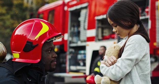 Porträt des geretteten kleinen asiatischen Mädchens mit dem Feuerwehrmannmann, der nahes Löschfahrzeug steht. Feuerwehrmann im Brandbekämpfungseinsatz