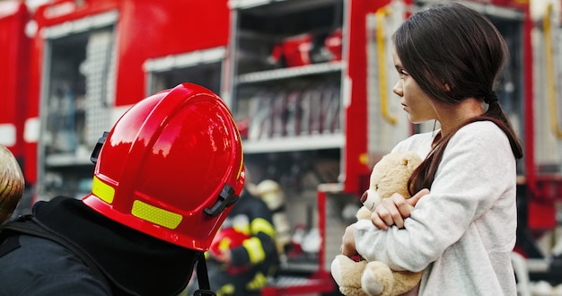 Porträt des geretteten kleinen asiatischen Mädchens mit dem Feuerwehrmannmann, der nahes Löschfahrzeug steht. Feuerwehrmann im Brandbekämpfungseinsatz