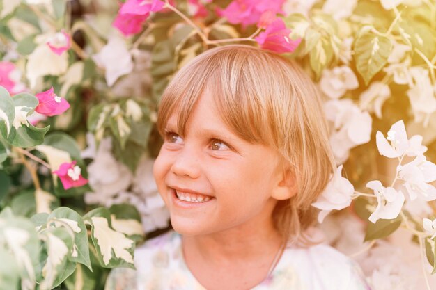 Porträt des Gesichts offener kleiner glücklicher lächelnder fünfjähriger blonder Junge mit grünen Augen in rosa und weißen Blumenpflanzen in der Natur Kinder haben Spaß an den Sommerferien helles Licht und luftiges Aufflackern