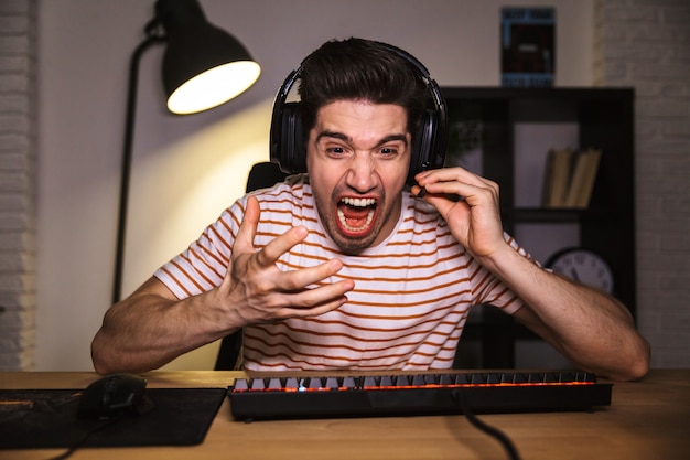 Porträt des gestressten jungen Mannes 20s, der schreiendes Headset trägt, während er am Schreibtisch im Raum sitzt und Videospiele spielt