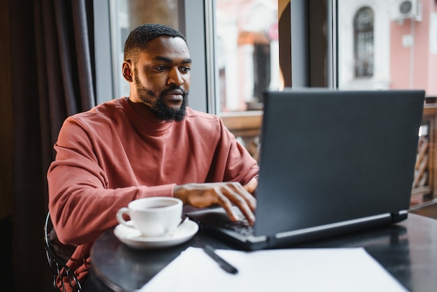 Porträt des glücklichen afrikanischen Geschäftsmannes, der in einem Café sitzt und am Laptop arbeitet