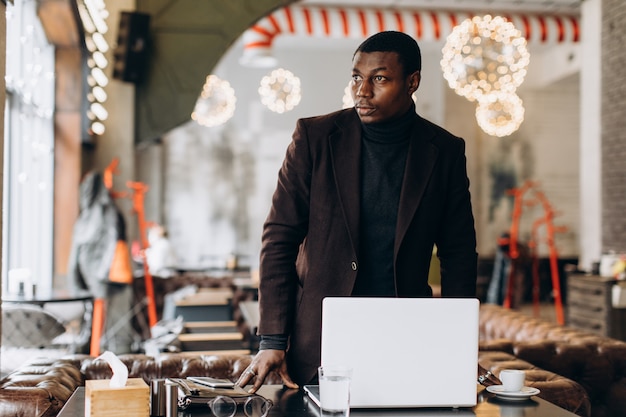 Porträt des glücklichen afrikanischen Geschäftsmannes unter Verwendung des Telefons beim Arbeiten an Laptop in einem Restaurant.