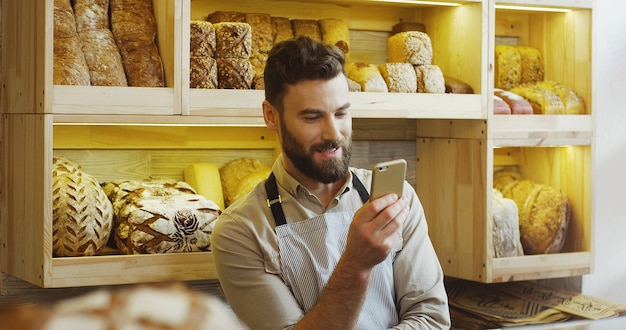 Porträt des glücklichen Bäckers, der im Bäckereigeschäft unter Verwendung des Smartphones arbeitet.