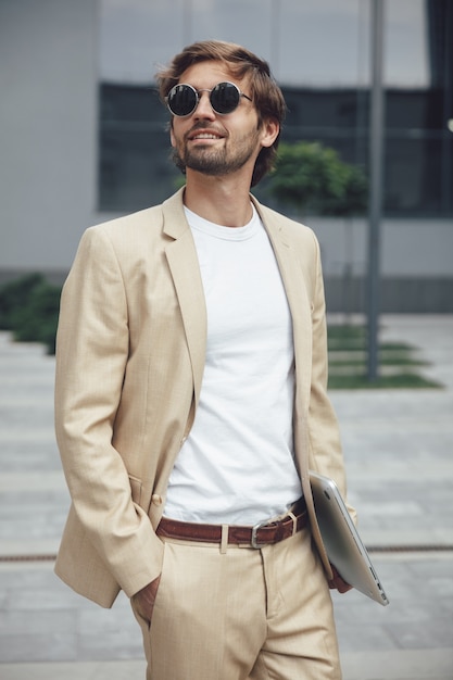 Porträt des glücklichen bärtigen Mannes in der formellen Kleidung und in der schwarzen Sonnenbrille, die drahtlosen Laptop draußen tragen