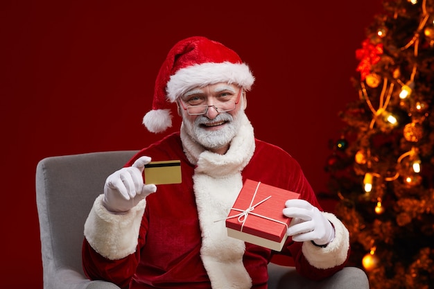 Porträt des glücklichen bärtigen Weihnachtsmanns, der Kreditkarte und Geschenkbox in seinen Händen hält, kauft er Weihnachtsgeschenke online