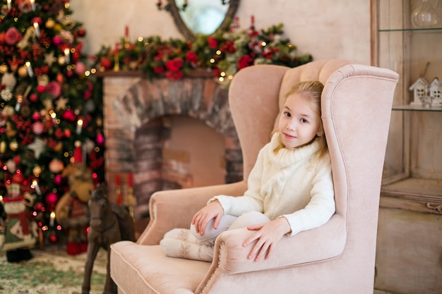 Porträt des glücklichen blonden Kindermädchens in der weißen Strickjacke, die auf dem Boden nahe dem Weihnachtsbaum stationiert