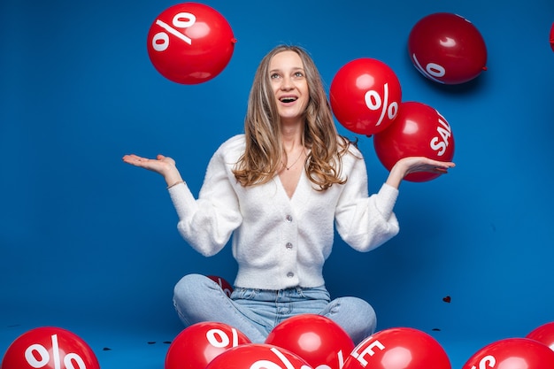 Porträt des glücklichen blonden Mädchens in der weißen Strickjacke und in den Jeans, die Arme halb gebogen halten und fliegende rote Luftballons mit Verkaufswort und Prozentzeichen betrachten