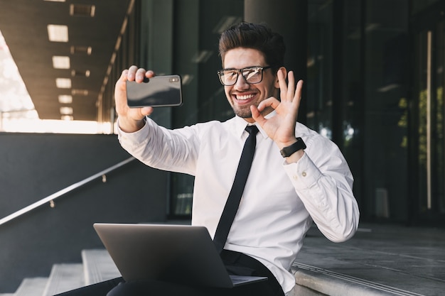 Porträt des glücklichen Geschäftsmannes gekleidet im formellen Anzug, der außerhalb des Glasgebäudes mit Laptop sitzt und Selfie-Foto auf Handy nimmt