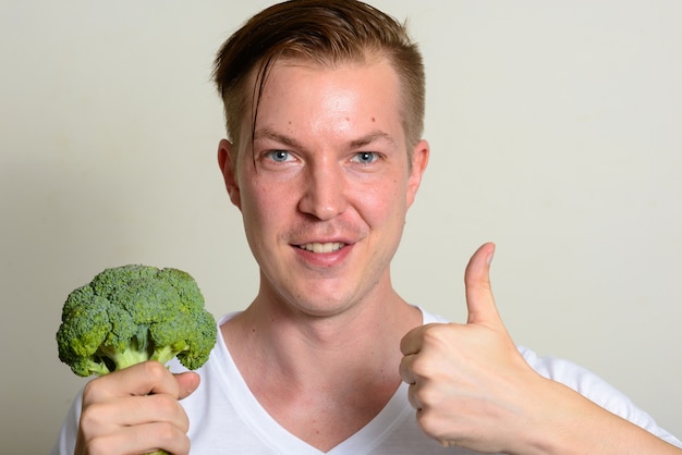 Porträt des glücklichen jungen gutaussehenden Mannes, der Brokkoli hält und Daumen aufgibt