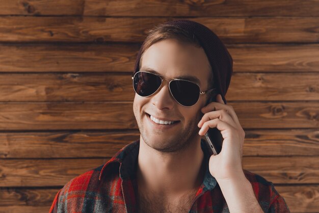 Porträt des glücklichen jungen Mannes im Hut und in den Spektakeln, die am Telefon sprechen