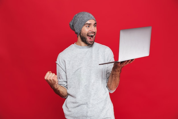 Porträt des glücklichen Kerls 30s in der Freizeitkleidung, die sich freut, während sie silbernen Laptop isoliert hält