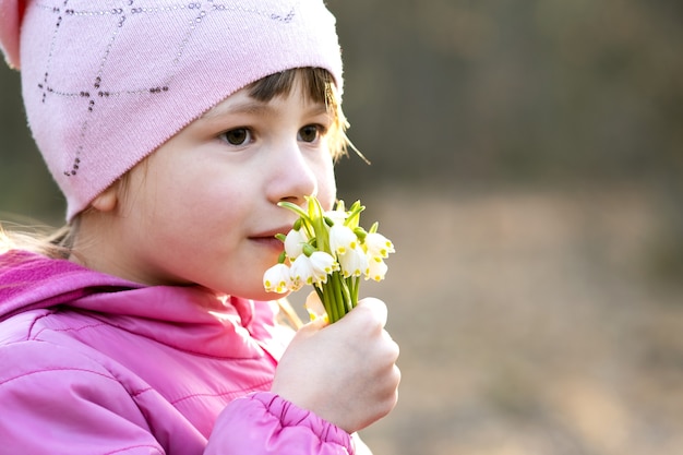 Porträt des glücklichen Kindermädchens, das Bündel der frühen Frühlingsschneeglöckchenblumen draußen hält.